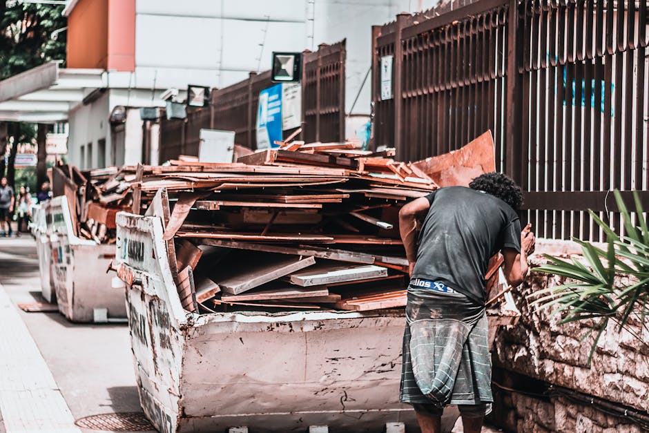 A man with curly hair wearing a black t-shirt and plaid shorts is standing on a city sidewalk next to a large white skip filled with wooden, tiled, and plastic waste. The skip contains a pile of disused roof tiles, broken wooden panels, and packaging materials, all stacked haphazardly. The skip is positioned next to a tall metal fence and nearby parked vehicles, with an urban environment visible in the background. This scene depicts the disposal of bulky waste following a home relocation, with the man possibly preparing for waste removal or loading materials into the skip. The setting is outdoors, under daylight, emphasizing the process of clearing debris associated with moving or renovation. The image relates to the services provided by Man with Van Abbey Wood, focusing on bulky waste removal and clearance in the context of house removals and moving logistics, supporting efficient transport and disposal of large household items.