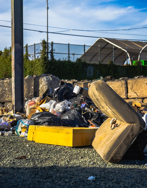 A pile of rubbish and discarded household items accumulated outside a property, including multiple black and white plastic garbage bags, scattered packaging materials, a yellow plastic box, an old, worn mattress leaning against a stone wall, and a wooden bed frame or furniture piece partially visible among the debris. The debris is situated on a gravel surface near the edge of a pavement, with a metal pole and overhead power lines in the background, alongside a green hedge, a large outdoor storage tent, and a blue sky with some clouds. This scene depicts waste collection after a home relocation or clearance, illustrating the sort of bulky waste removal services offered by Man with Van Abbey Wood, supporting efficient and responsible cleanup and disposal following a move or renovation.