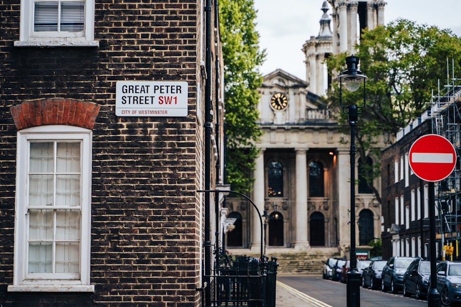 A street scene in Abbey Wood, showing a brick building with a white-framed window and a street sign that reads 'Great Peter Street SW1, City of Westminster,' positioned on the corner of the building. In the background, a historical building with classical architecture, including columns and a clock, is visible, partially obscured by green trees. A black street lamp is situated along the sidewalk, and several parked cars line the side of the street. A red and white 'no entry' traffic sign is placed on the right side of the image. The scene captures the urban environment where house removals and furniture transport often take place, with evidence of temporary scaffolding on adjacent buildings and a clean, well-maintained pavement area, reflecting typical moving logistics in central London, associated with services like those provided by Man with Van Abbey Wood.