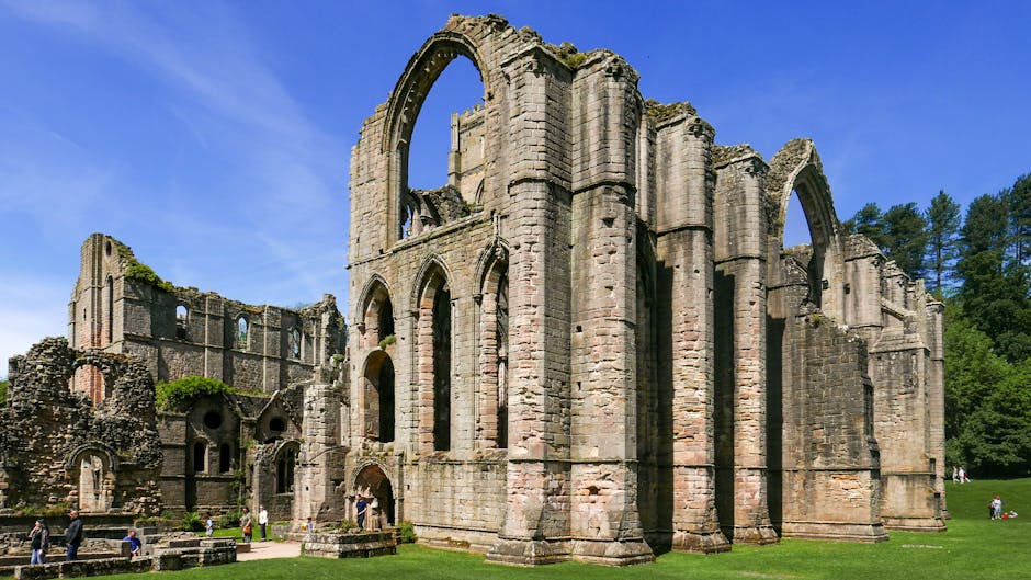 A large, weathered stone building with tall, pointed arches and multiple windows, some of which are missing glass or completely open. The structure appears to be a historic ruin with sections of the walls and roof absent, exposing the sky above. In the foreground, a paved pathway is visible, with several people walking or standing nearby, some taking photographs. Surrounding the ruins is a well-maintained grassy area with a few scattered trees, and a clear blue sky with a few wispy clouds overhead. The scene suggests a site of historical significance, and the image captures the intricacies of its stonework, including columns and buttresses typical of medieval architecture. This setting is associated with house removals and packing and moving services, highlighting the importance of careful transportation of antiques, artwork, or historical items during a home relocation, as referenced on the Man with Van Abbey Wood website.