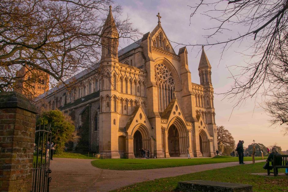 A large, historic Gothic-style church building constructed from light-colored stone, featuring intricate architectural details such as pointed arches, large stained glass windows, and tall spires. The church is situated outdoors on a paved pathway, with leafless trees on either side and a grass lawn surrounding it. Several people are visible, some standing near the entrance and others walking along the pathway, with a black metal gate and brick pillar partially visible in the foreground. The scene appears to be captured during late afternoon or early evening with a soft pinkish-purple sky, highlighting the building’s detailed facade. This setting reflects the context of house and furniture removals, as the area might be used for loading or unloading during a home relocation process. [COMPANY_NAME], experienced in removals and moving logistics, may utilize similar environments during furniture transport and packing activities, ensuring safe and efficient home relocation services near Lesnes Abbey.
