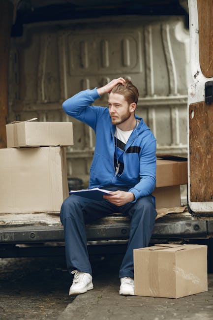 A man dressed in a blue long-sleeve shirt, black vest, black trousers, and white sneakers stands outside a residential property next to a white moving van with a wooden panel on its side. He is wearin