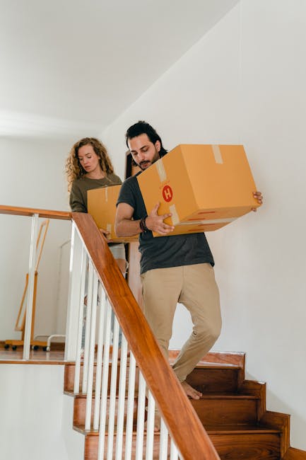 A man and woman are carrying cardboard boxes as part of a home relocation process inside a residential property, with the man holding a box in front while descending wooden staircase steps, and the woman following behind with another box. The staircase has white spindles and a wooden handrail, and the interior space is well-lit with natural light. The boxes are sealed with packing tape, and the scene captures the manual handling aspect of furniture transport and packing during house removals. This action takes place on a landing area at the top of the stairs, with visible wooden flooring and plain white walls. The presence of packing materials and careful lifting reflects the systematic moving service offered by Man with Van Abbey Wood, supporting efficient and secure furniture transport within the house clearance process.