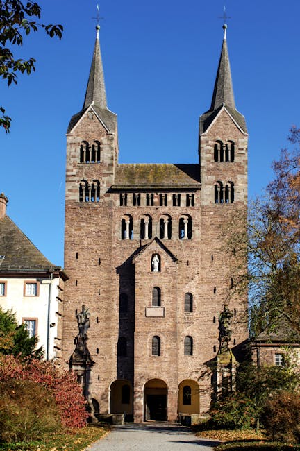 A tall, historic stone church with two pointed spires, each topped with a weather vane, stands prominently against a clear blue sky. The church features narrow, elongated windows with rounded arches, arranged in symmetrical tiers on its façade, and a central entrance with a small gabled roof. The building is surrounded by trees and bushes, some with autumn foliage, and a gravel pathway leads up to the entrance. The scene captures the exterior of the church during daylight hours, with natural light illuminating the stonework and casting shadows. This image relates to house removals and moving services provided by Man with Van Abbey Wood, emphasizing the importance of careful transport and handling of varied structures and belongings during relocations, although in this case, it depicts a building rather than furniture or boxes.