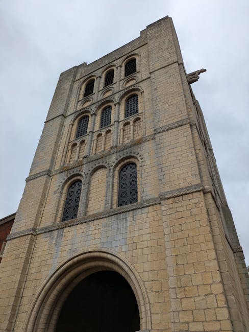 Photograph of a large, historic stone church tower with multiple levels of arched windows framed by decorative stonework, viewed from ground level against an overcast sky. The tower's beige and light brown stone façade shows signs of weathering. The arched entrance at the base is partially visible, with chipped stone details around it. The tower is situated on a paved area, with no visible vehicles or people nearby. This structure exemplifies traditional ecclesiastical architecture and features a crucifix sculpture extending from the side of the tower near the top. The image emphasizes the height and intricate masonry of the building, suitable for illustrating house removals or building access in the context of relocation or packing and moving processes, as provided by Man with Van Abbey Wood.