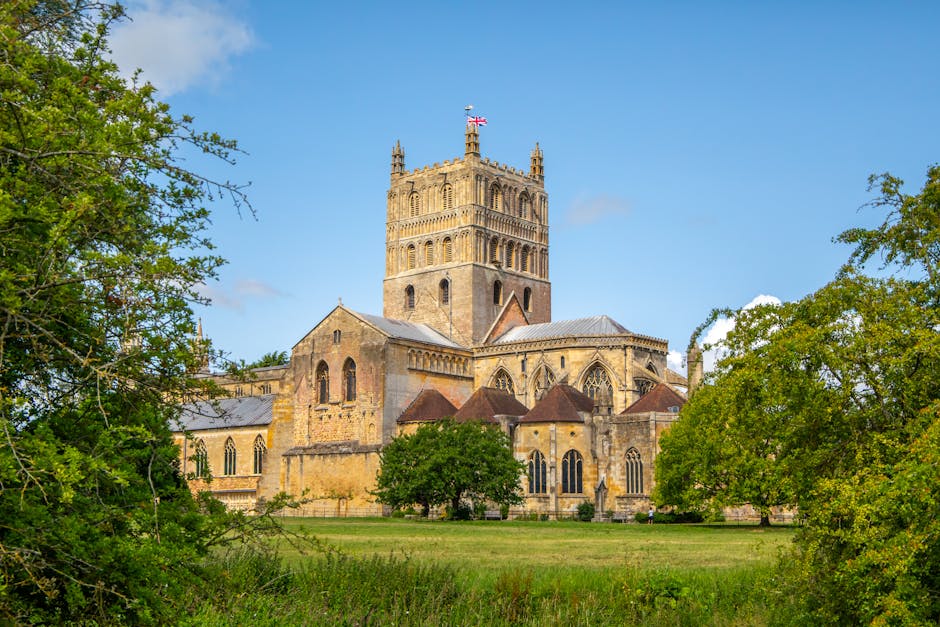 The image shows a historic stone church with a tall central tower topped by a small spire and a Union Jack flag flying at the peak, set against a bright blue sky with scattered clouds. Surrounding the church are lush green trees and well-maintained grass, with some smaller buildings and structures attached to the main building. The scene is captured during daylight, with natural lighting highlighting the detailed architectural features such as arched windows, buttresses, and decorative stonework. This setting could depict a typical location relevant to house removals or relocation services, emphasizing the significance of careful planning during home relocation processes. Man with Van Abbey Wood might utilize such imagery to illustrate their comprehensive moving logistics, including packing, furniture transport, and loading procedures in a residential or historic context.