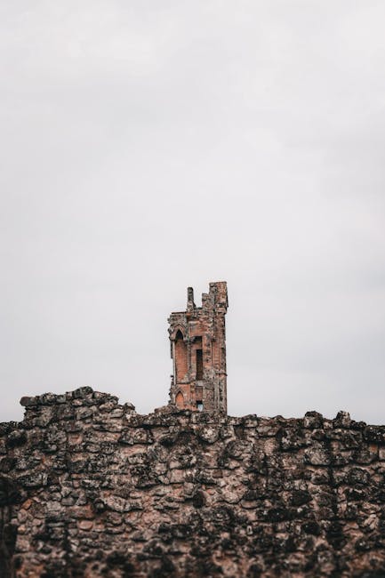 Photograph of a large, historic stone church tower with multiple levels of arched windows framed by decorative stonework, viewed from ground level against an overcast sky. The tower's beige and light brown stone façade shows signs of weathering. The arched entrance at the base is partially visible, with chipped stone details around it. The tower is situated on a paved area, with no visible vehicles or people nearby. This structure exemplifies traditional ecclesiastical architecture and features a crucifix sculpture extending from the side of the tower near the top. The image emphasizes the height and intricate masonry of the building, suitable for illustrating house removals or building access in the context of relocation or packing and moving processes, as provided by Man with Van Abbey Wood.
